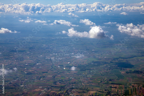 An aerial view from an airplane window looking down at fields, towns, and scattered cloud formations under a bright blue sky. The landscape below displays a patchwork of agricultural fields and roads.