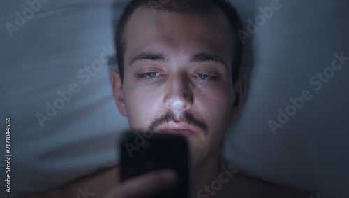 Fotografie Young Man Lying in Bed at Night Using Smartphone in Dark Room