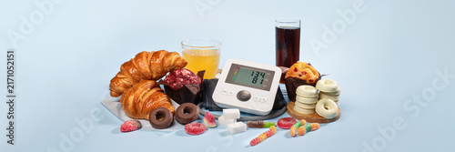Blood pressure monitor surrounded by unhealthy foods: croissants, donuts, muffins, chocolate, gummies and sweet drinks. Hypertension and cardiovascular risk diet concept, web banner, copy space