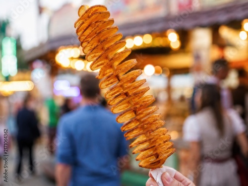 Knusprige Spiralkartoffel am Holzspieß, beliebter Snack auf Volksfest und Jahrmarkt. Goldbraun gewürzt und vor einem belebten Verkaufsstand mit warmen Lichtern fotografiert.