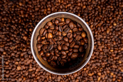 Top view of metal grinder container filled with roasted coffee beans on coffee bean texture background for espresso preparation, specialty brewing, and cafe concept.