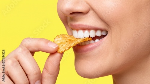 Close up young woman biting crispy potato chip against bright yellow background for snack food advertisement with view person