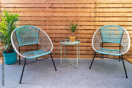 Modern home terrace with wooden slate screen, exterior carpet, retro style grey and turquoise chairs, small round metal table and plants 