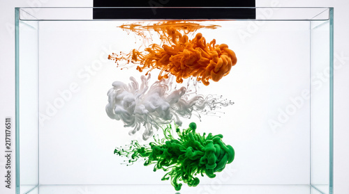 Vibrant orange, white, and green liquid paint suspended and flowing inside a transparent glass tank, creating an abstract artistic composition with colorful fluid movement.