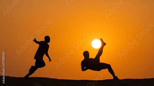Silhouette of a man kicking the sun while another runs on a sand dune with running athlete concept with sunset