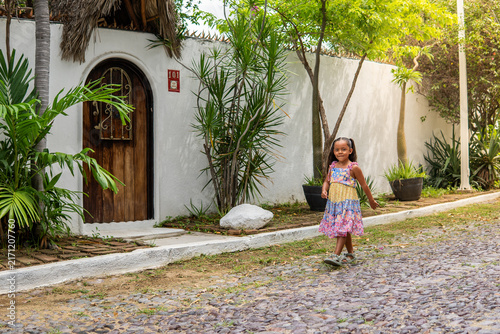 niña morena latina feliz con vestido colorido caminando por una calle empedrada pintoresca afuera de una casa de playa con plantas y palmeras verdes disfrutando de un hermoso dia soleado en el mar 