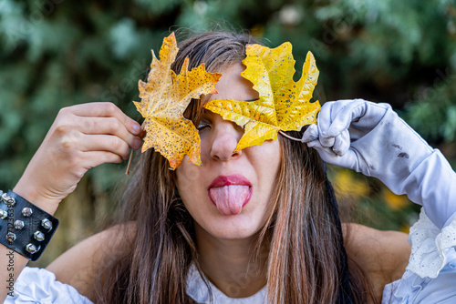 Playful woman posing with yellow autumn leaves covering her face while sticking out her tongue in a forest, wearing a white dress, gloves and a black studded bracelet.