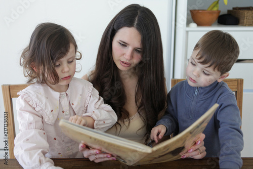 Fotografie A mother reads a storybook with her young children at a wooden table at home, encouraging early learning, literacy, family bonding and quality time together