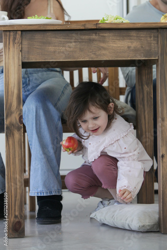 Obraz na płótnie Playful little girl hiding and playing under the dining table during a family meal at home, holding an apple and enjoying a happy childhood moment