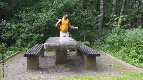Young woman sitting on a wooden bench in a peaceful forest park, wearing a yellow T-shirt and white jeans. Natural outdoor lifestyle scene surrounded by green trees and summer vegetation.