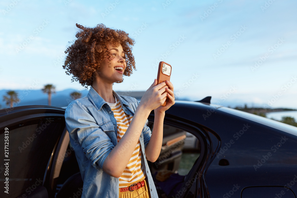 Naklejka premium Cheerful woman outdoors, smiling while holding a phone, beside a car, sunny coastal scene, denim shirt, striped top, relaxed lifestyle, natural colors, soft breeze, warm light, palm trees.