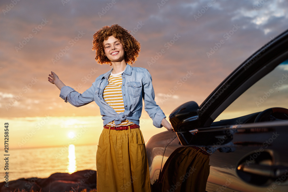 Naklejka premium Young woman with curly hair smiles beside a car at sunset, wearing a denim jacket over a striped top and a yellow skirt, embodying a carefree lifestyle and warm golden light.