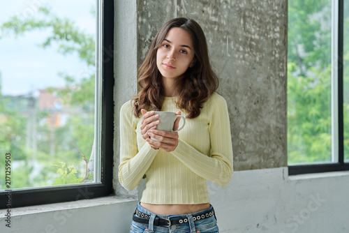 Young attractive woman holding coffee mug standing near window looking at camera