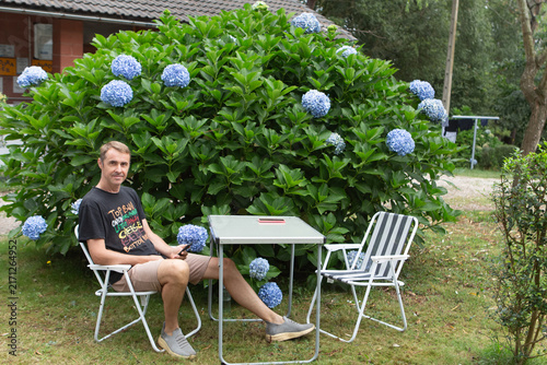 Man sits at a table in front of a large bush with blue flowers, enjoying a moment outdoors on a sunny day
