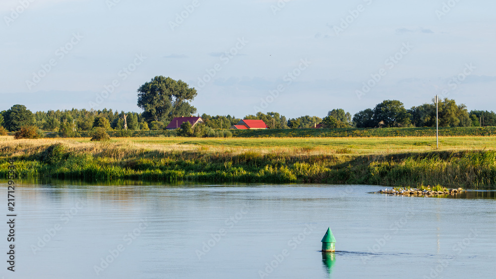 Obraz premium A green navigation buoy floats in a calm river with village houses and green meadows under a blue sky in summer.