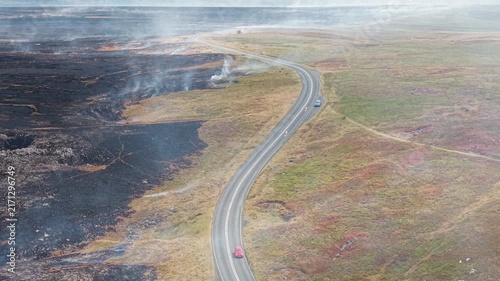 Red car moving along country road through burnt moorland, smoke drifting over burnt heather and grass after hill fire in Brecon Beacons National Park