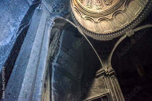 Dramatic stone arches frame an ornate, carved stalactite dome inside Geghard Monastery. Shafts of natural light pierce the dim, ancient chamber, highlighting medieval Armenian architecture