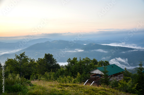 Wooden shelter with a green roof sits on a grassy hillside overlooking misty mountain peaks. Low clouds drift through the valleys under a soft sunrise sky, creating a serene alpine view