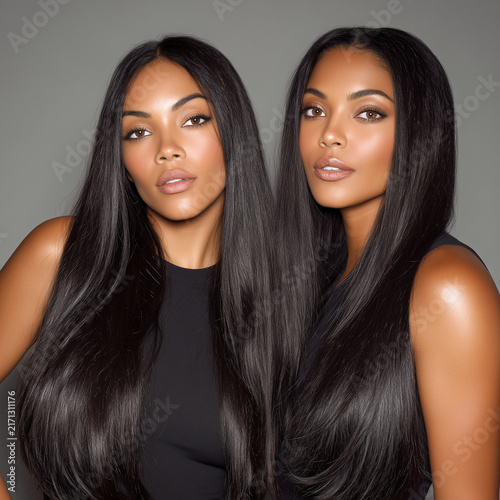 Two beautiful twin women with long straight shiny dark hair on gray background. Multiethnic female models with glowing makeup in black tops posing together. Beauty care concept