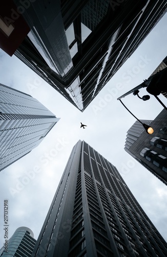 Quadro su tela Low angle view of airplane flying above modern skyscrapers converging in financi
