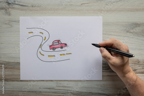 Photo of hand drawn graphic of red vehicle driving on a wavy road birds eye view with hand holding a black ink pen. Traveling road trip concept.