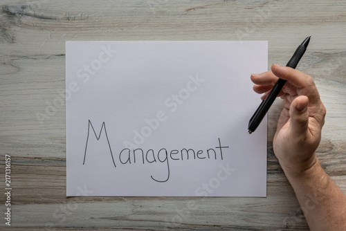 Flat lay background white sheet of paper with negative space available with the word Management handwritten on it with a hand holding a black pen.