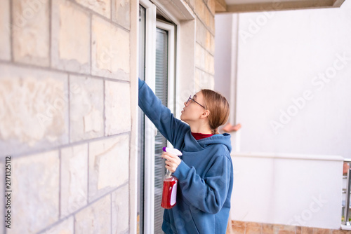 Teenage girl cleaning a balcony window with spray and cloth.