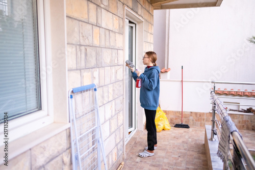 Teenage girl cleaning a balcony window with spray and cloth.