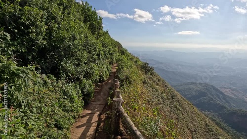A wooden plank trail runs along a cliff edge surrounded by trees and bushes, overlooking lush green hills and mountains in northern Thailand on a sunny day.