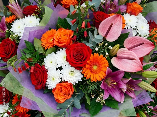 A vibrant and colorful bouquet of assorted flowers, including roses, lilies, gerbera daisies, and anthuriums, arranged with lush greenery