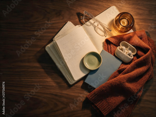 Warm light glows softly over a cozy wooden table. Open notebook rests beside glasses and a candle. Earbuds and a bowl sit nestled in a knitted scarf. Soft tones evoke comfort, quiet reflection