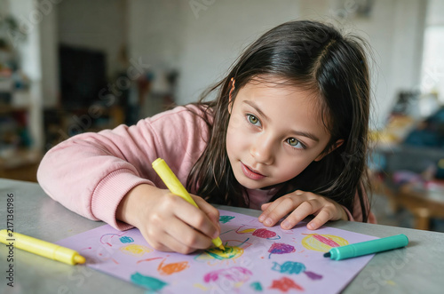 Young girl draws with colorful crayons. Focused eyes show quiet concentration. Soft pink sleeves hug her small arms. Crayon strokes create playful fish shapes. Background hints at cozy, lived-in space