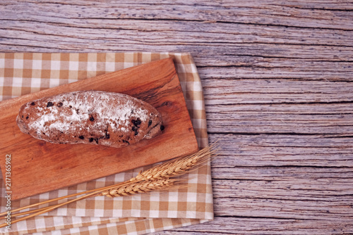Fresh sourdough rests on wooden board, dusted with flour. Warm bread sits atop rustic cutting board. Wheat stalks lie beside it, woven mat below. Subtle shadow hints at soft, cozy kitchen light