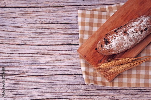 Fresh sourdough rests on wooden board, dusted with flour. Warm bread sits atop rustic cutting board. Wheat stalks lie beside it, woven mat below. Subtle shadow hints at soft, cozy kitchen light
