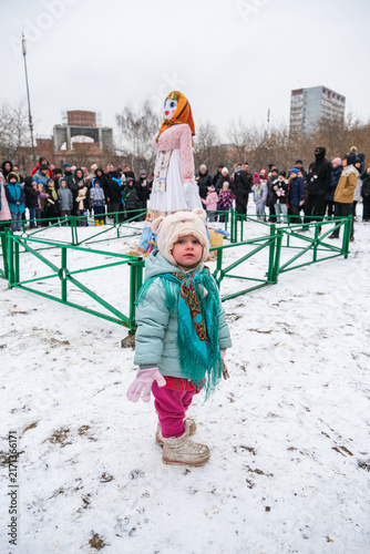 Quadro su tela A little girl standing on a snowy ground during a folk festival with a large Maslenitsa doll in the background