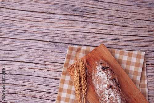 Fresh sourdough rests on wooden board, dusted with flour. Warm bread sits atop rustic cutting board. Wheat stalks lie beside it, woven mat below. Subtle shadow hints at soft, cozy kitchen light