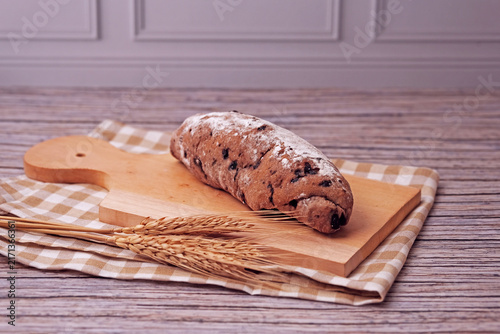 Fresh sourdough rests on wooden board, dusted with flour. Warm bread sits atop rustic cutting board. Wheat stalks lie beside it, woven mat below. Subtle shadow hints at soft, cozy kitchen light