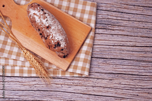 Fresh sourdough rests on wooden board, dusted with flour. Warm bread sits atop rustic cutting board. Wheat stalks lie beside it, woven mat below. Subtle shadow hints at soft, cozy kitchen light