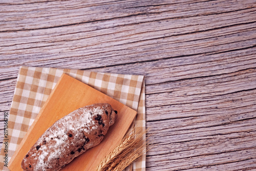 Fresh sourdough rests on wooden board, dusted with flour. Warm bread sits atop rustic cutting board. Wheat stalks lie beside it, woven mat below. Subtle shadow hints at soft, cozy kitchen light