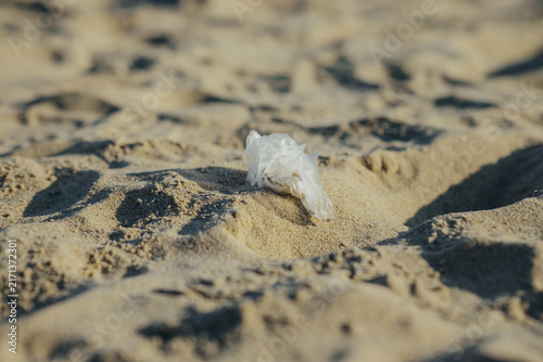 Crumpled plastic bag lying on sandy beach with footprints. Plastic waste pollution on seashore. Environmental problem of ocean litter and marine debris. Ecological conservation concept.