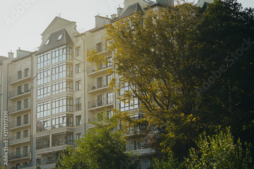 Modern residential apartment building with balconies behind green trees. Luxury housing complex facade view from below. Real estate and urban architecture concept.