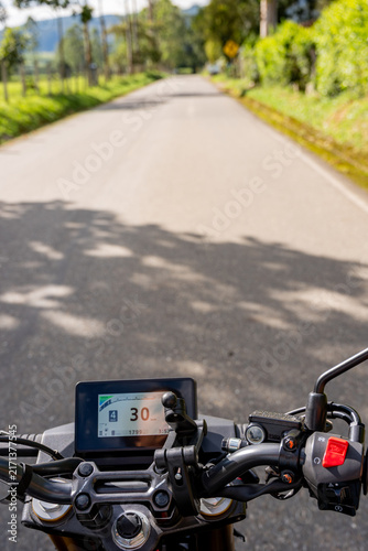 Motorcycle rider's point of view on a rural road, with handlebars and digital dashboard showing speed, enjoying freedom and adventure during a journey in the countryside