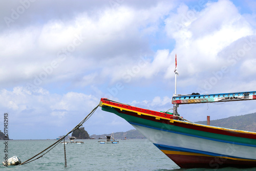 A colorful wooden fishing boat anchors in calm coastal waters beneath a cloudy blue sky