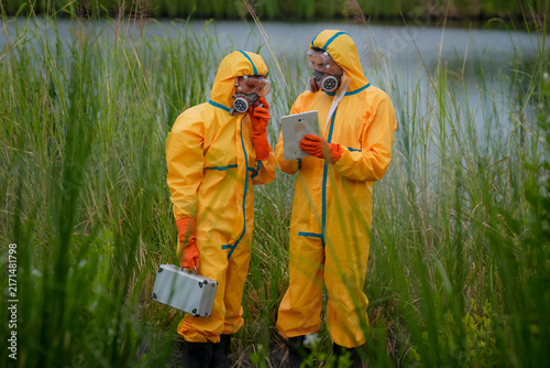 Hazard suit respirator environmental testing wetland scientist tablet water pollution fieldwork, team in protective clothing collecting sample near river reeds, focused and cautious