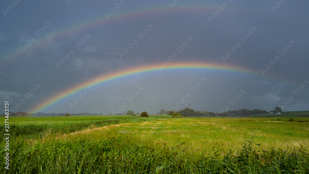 Naklejka premium Rainbow arches across green field. Colorful rainbow follows dramatic weather. Rainbow enhances rural landscape. Brilliant rainbow dominates cloudscape naturally.