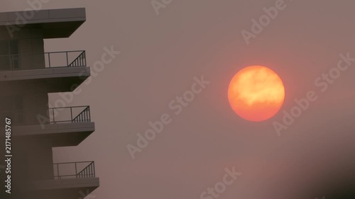 Smoke from forest fire fills the sky, leaving the sun in a deep orange glow in a city with high rise apartment building.