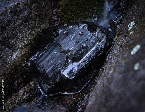 Obsidian Stone Wedged in a Moisture-Seeping Rock Crevice, Water Film and Droplets Covering Its Striated Volcanic Glass Surface