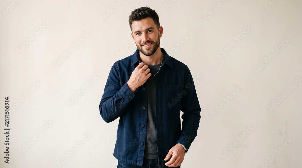 Obraz premium Portrait of a handsome young man with a beard wearing a dark blue overshirt and grey tshirt, smiling and looking at the camera against a neutral background