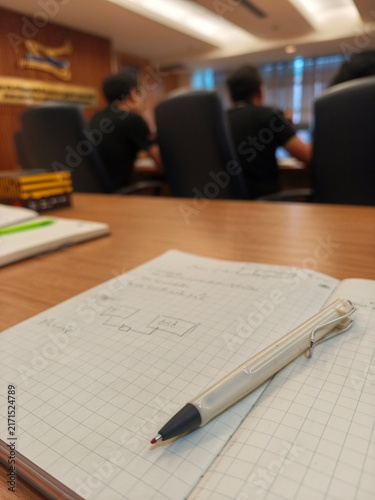 A low-angle close-up shot of an open grid-paper notebook with a pen resting on it, placed on a wooden conference table with a shallow depth of field, blurring people sitting in a meeting room 