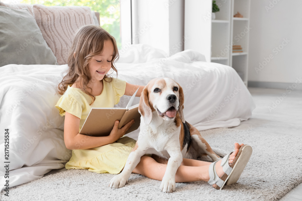 Fototapeta premium Cute little girl with Beagle dog reading book on floor in bedroom